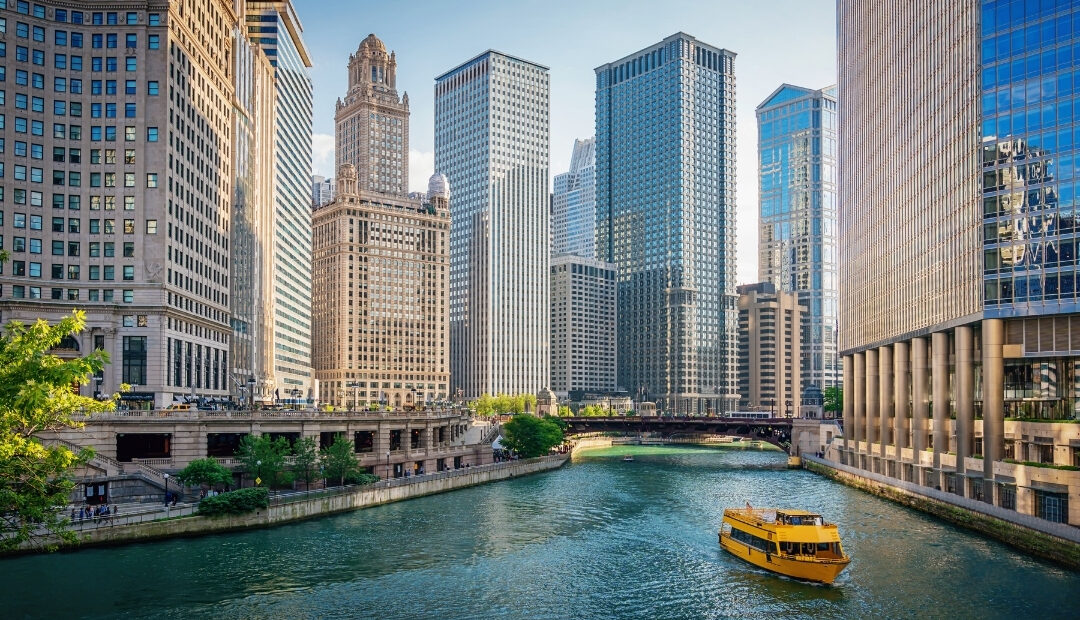 A scenic view of Chicago's skyscrapers lining the Chicago River, with a yellow architecture tour boat passing through. Book your bus tickets to Chicago.
